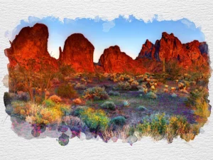 Fiery red rock formations rise above vibrant desert flora in fading sunlight.