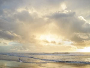Golden sunlight breaks through clouds above calm waves, illuminating the tranquil ocean horizon.