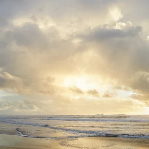 Golden sunlight breaks through clouds above calm waves, illuminating the tranquil ocean horizon.