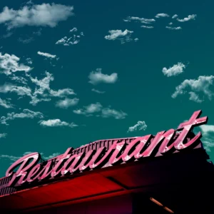 The pink sign of the restaurant sits in bright contrast to a teal sky dotted with clouds.