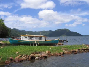 A cloudy sky shades deep green mountains in the background of a bright teal and white boat, run aground at a dock.