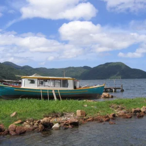 A cloudy sky shades deep green mountains in the background of a bright teal and white boat, run aground at a dock.