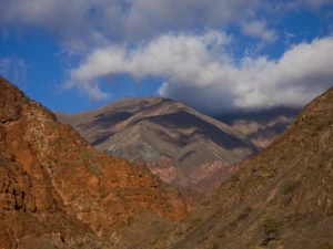 Red craggy mountains shield a neighboring mount that is engulfed in approaching clouds under a vast blue sky.