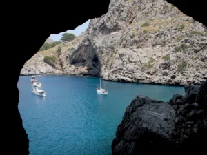 Peaking from a sea cave, leisure boats drift into view over aquamarine water, backed by rocky, limestone cliffs.