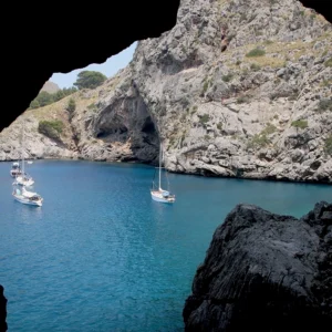 Peaking from a sea cave, leisure boats drift into view over aquamarine water, backed by rocky, limestone cliffs.