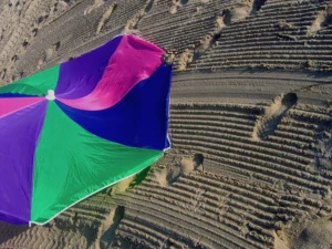 A partially collapsed multi-colored umbrella lay discarded on a popular sandy beach littered with various footprints and tire treads.