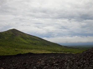 Dark rocky gravel litters the foreground of a sprawling green valley that climbs into a steep peak beneath a boundless cloudy sky.